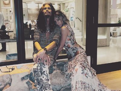 Steven Tyler is sitting with his legs folded while Aimee Preston is leaning into him.